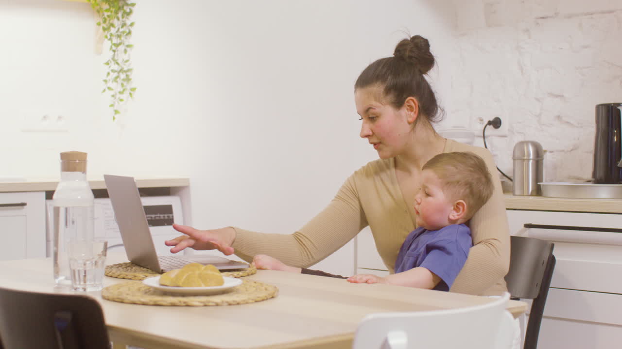 Young Mother Working With Laptop Computer While Holding Her Baby Boy At Home