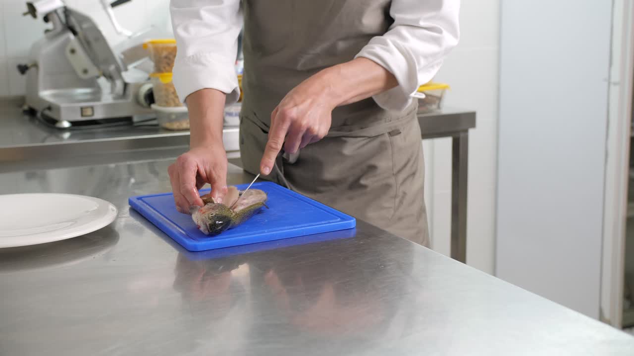 Male cook fillets raw fish with knife in kitchen, medium pan shot