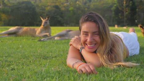 hermosa chica riendo mientras está acostada sobre la hierba verde con la barbilla apoyada en los brazos - chica con canguros grises del este - costa dorada, qld, australia