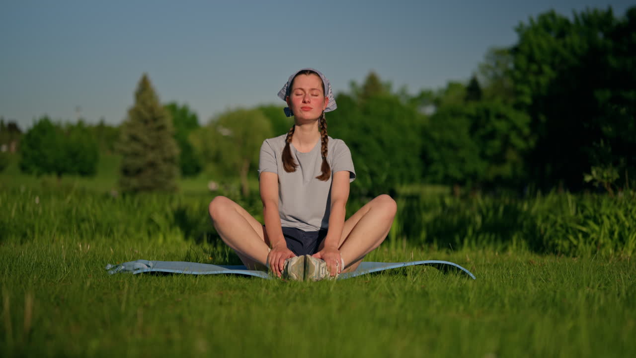 A young woman settles onto her yoga mat and smiles, embracing the beautiful sunlight and the positive energy of nature. She enjoys this peaceful outdoor moment. Gentle left travelling.