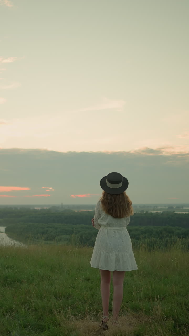 A woman in a white dress and hat stands with folded arms, thoughtfully in a grassy field overlooking a tranquil lake at sunset. She gazes at the peaceful environment