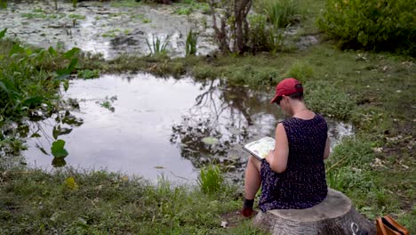 camera moves up and around a woman in a dress sitting beside a stump drawing and painting in a book beside a pond