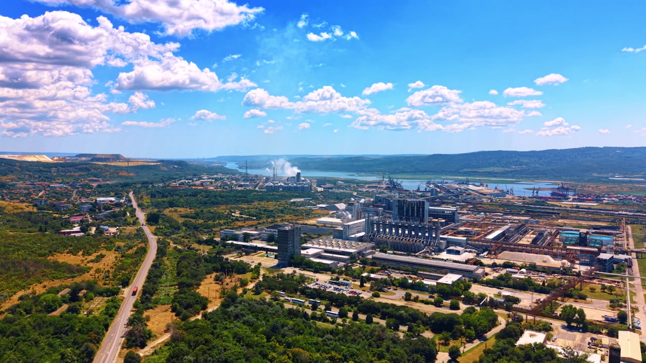 Ampio paesaggio con strada che porta verso una zona industriale. Fogliame verde e campi aperti costeggiano un'autostrada asfaltata sotto un cielo azzurro parzialmente nuvoloso