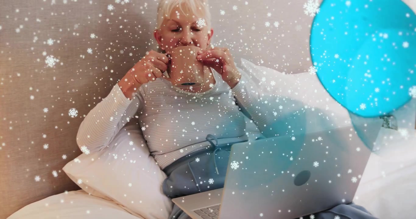 Mulher idosa pegando caneca com flocos de neve e pontos azuis, bebendo perto do laptop para bem-estar em casa.