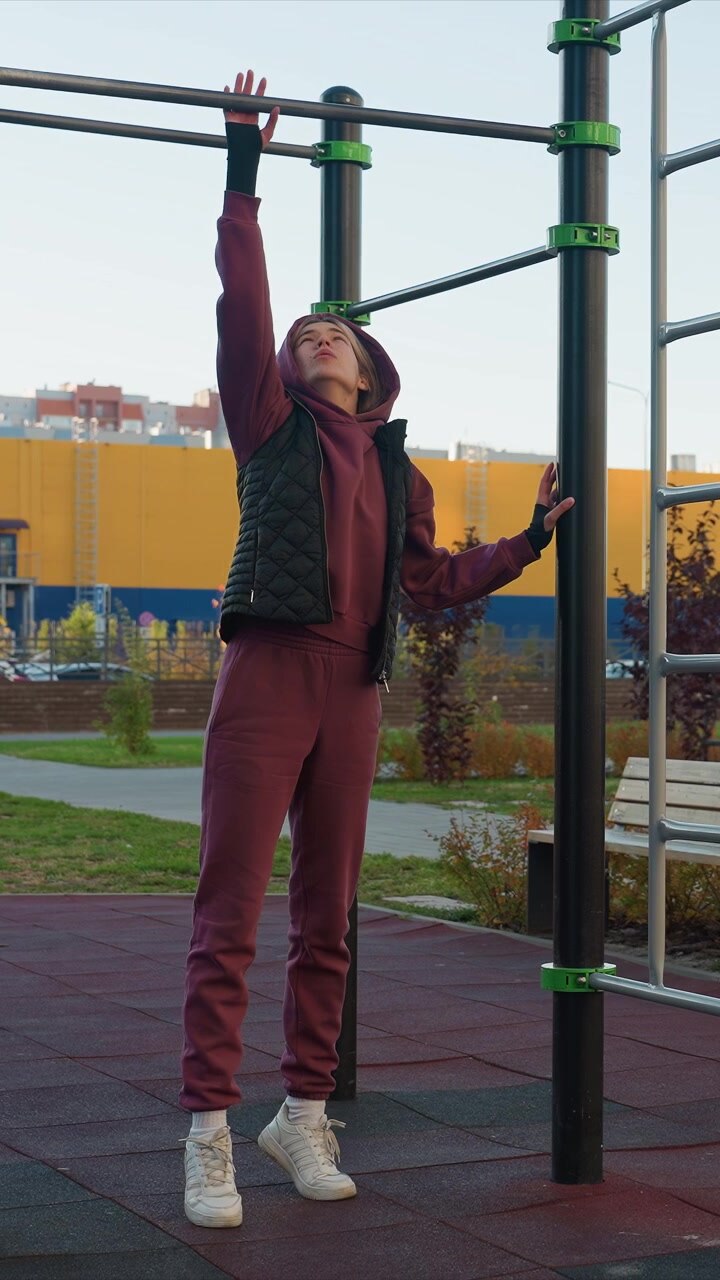 Woman exercises outdoors using playground bars in urban courtyard. Woman stretches and prepares for bodyweight movements wearing maroon tracksuit with black vest against backdrop of city