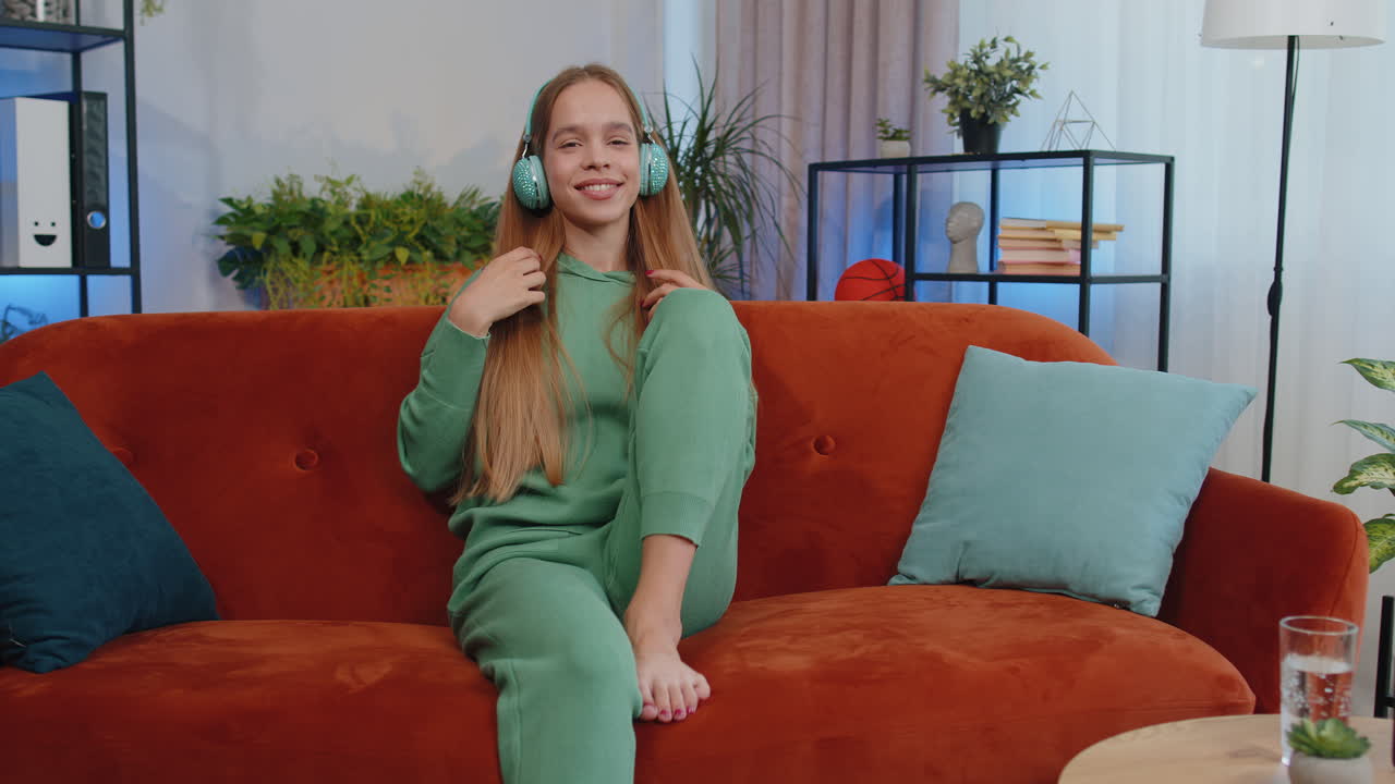 무선 헤드폰을 입은 행복하고 편안한 소녀가 춤을 추고 집 소파에서 좋아하는 록 앤 롤 음악을 듣고 있습니다.