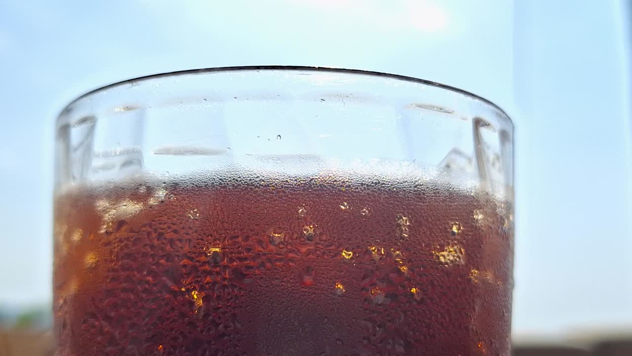 Close-up of cold Coke in a glass with bubbles, blue sky and sun in the background