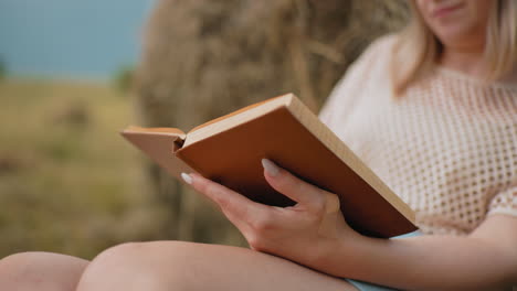 primer plano de una mujer volviendo a una nueva página mientras lee al aire libre contra una bala de heno, las manos sujetan suavemente el libro, revelando páginas alineadas, con un borrón suave en el fondo