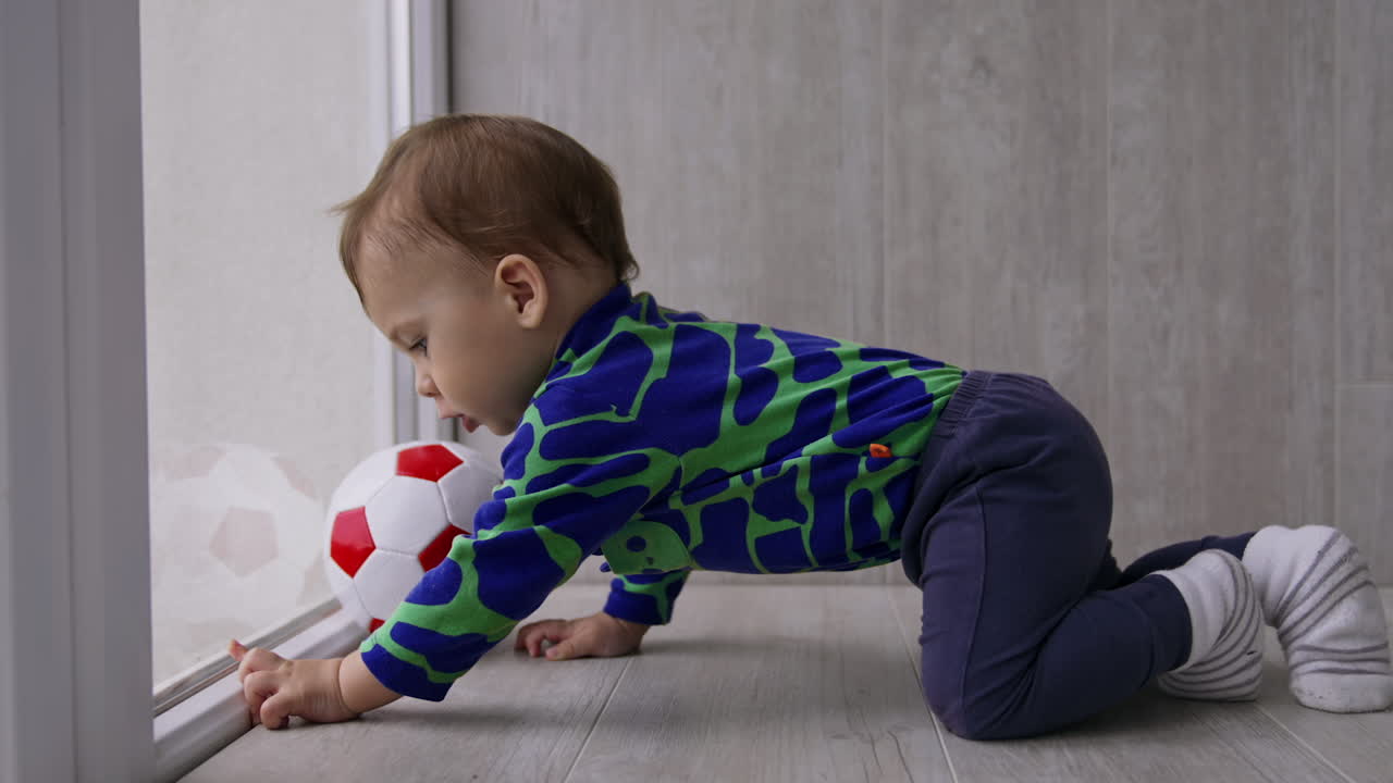 Toddler boy crawling up to the ball near the window. Cute kid moves the ball and follows it. Side view.