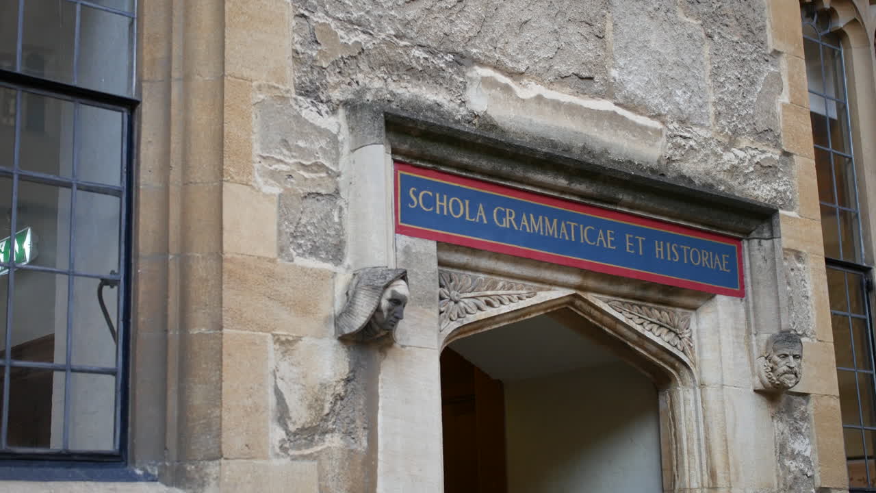 Døråpning ved Oxford University med latinsk inskripsjon 'school of grammar and history' over en utskåret steinbue, omgitt av forvitret murverk og blyglassvinduer