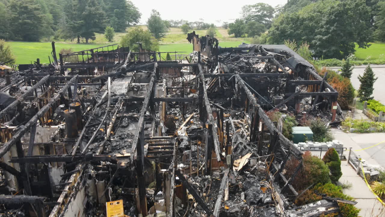 드론이 천천히 올라가는 모습. 앞면에는 불타버린 건물이 있고, 뒤에는 멀리서 산이 있는 호수 연안이 보인다.