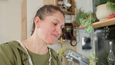 lady in green apron smiles warmly while showcasing thick mixed flour in cozy kitchen, surrounded by rustic tools, potted plants, and electric mixer