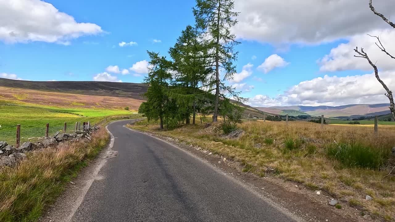 Car drives along winding rural road through scenic Scottish countryside under partly cloudy summer skies