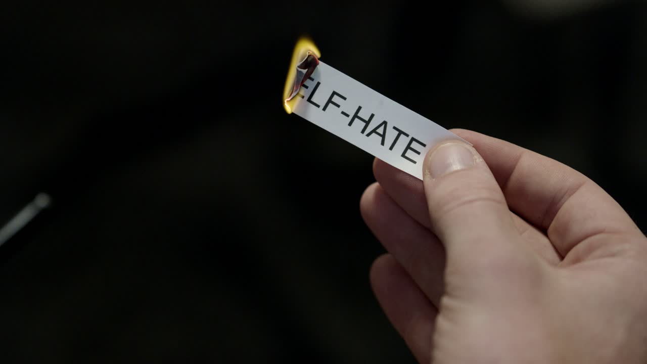 Eliminare l'odio verso se stessi - Mano che dà fuoco a un biglietto di carta con la scritta “ODIO VERSO SE STESSI”, potente liberazione da critiche, sensi di colpa e vergogna, primo piano cinematografico scuro. Salute mentale, terapia