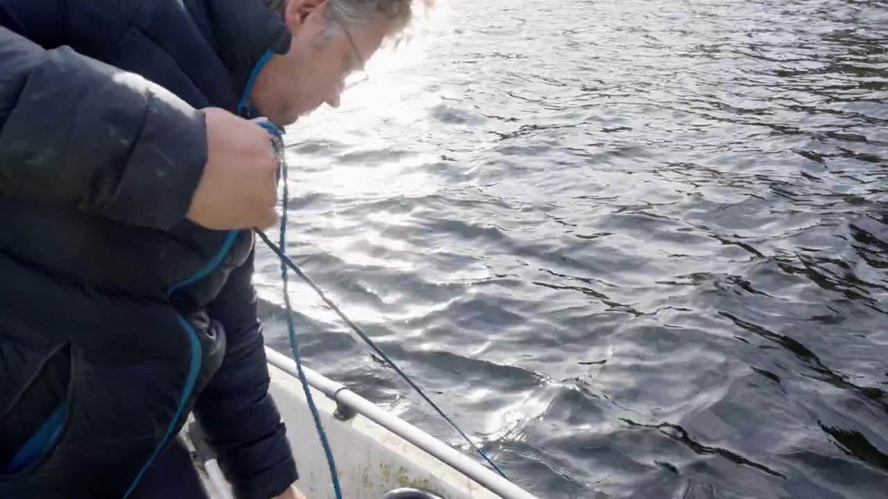 Un uomo sulla quarantina con i capelli ricci solleva una rete per granchi dal mare nella barca, ripresa a mano con l'acqua increspata sotto.