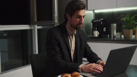 A-serious-brunette-man-with-stubble-in-a-black-suit-sits-at-a-table-in-front-of-a-laptop-and-communicates-via-video-conference-while-sitting-without-pants-only-in-shorts-in-a-modern-kitchen-in-the-evening-at-home