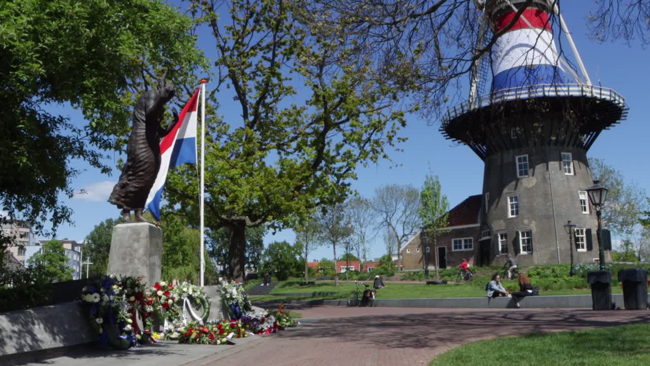 Flowers and wreaths laid out at World War Two monument in the Netherlands, flanked by flag post with Dutch flag, and with flag-wrapped windmill in background