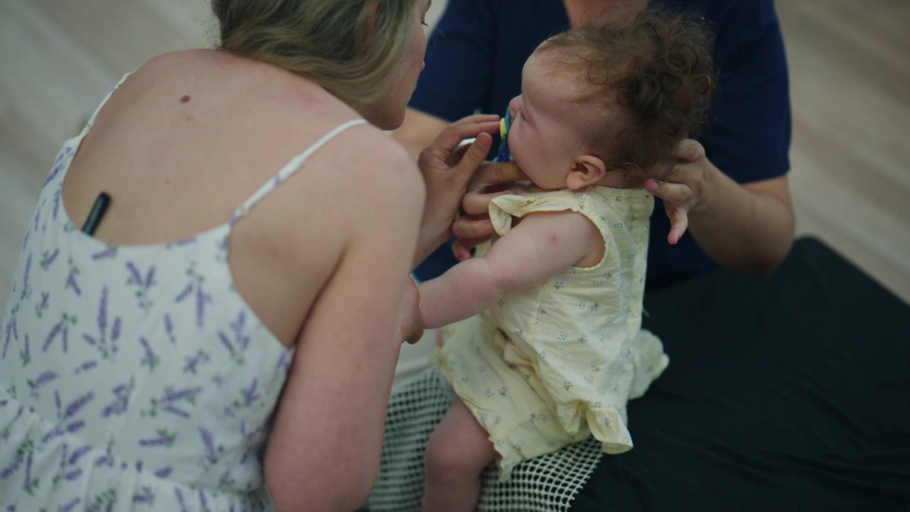 Baby ruht mit Schnuller auf dem Schoß eines Erwachsenen, während eine andere Person sanft Kopf und Körper während einer ruhigen Pflegeroutine stützt.