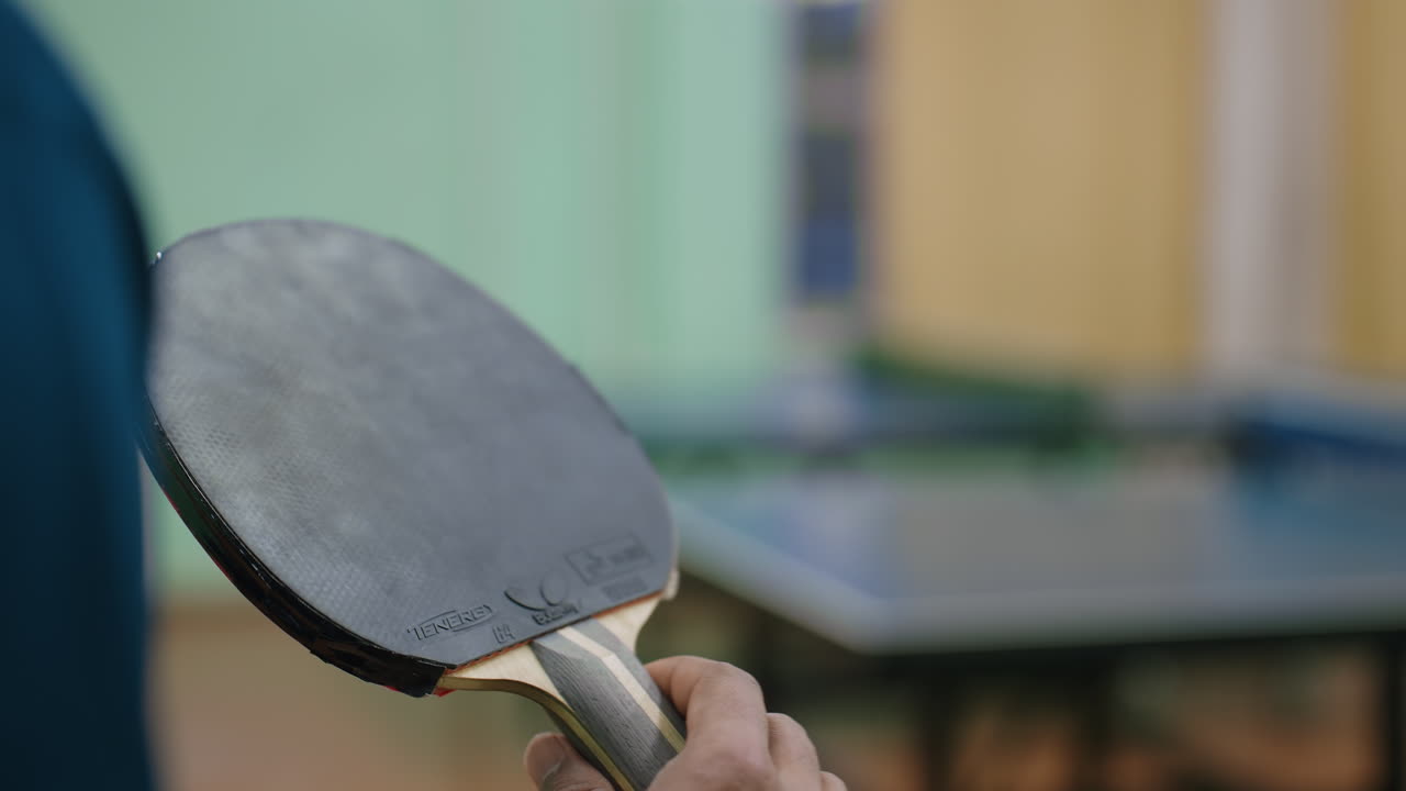 Nærbilde av hånd som holder tennisracket mens den snur under spill, fokus på detaljer på racketen med en uskarp innendørs bakgrunn som viser et subtilt spillmiljø