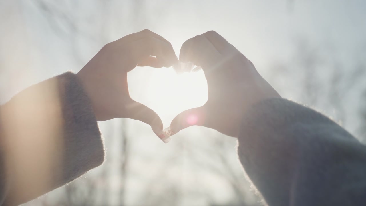 Belle mani di donna bianca che formano un cuore, primo piano intimo retroilluminato da un sole forte, manica del cappotto visibile e bagliore ad aureola, toni caldi da pellicola, modella in posa per il fotografo, movimento tenero e spontaneo.