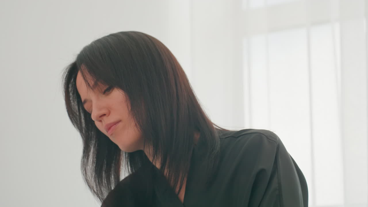 明るいサロンで髪をチェックするヘアスタイリスト、黒いスモックを着た女性が穏やかで集中的な表情で頭を傾げている、薄手のカーテンから差し込む柔らかな自然光、頭と肩のクローズアップ、評価するスタイリスト。