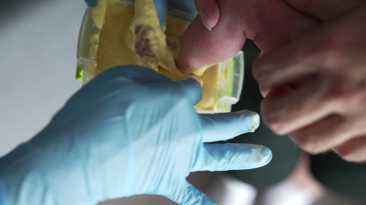 Tiny baby foot is put into the yellow stuff in the container. Hands in latex gloves holding the kid's foot. Close up.