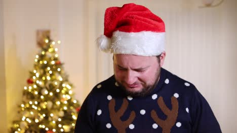 Triste-Hombre-Caucásico-Barbudo-Con-Sombrero-De-Santa-Llorando-Solo-En-Su-Casa-Con-árbol-De-Navidad-En-El-Fondo,-Depresión-Soledad-Durante-Las-Vacaciones-De-Navidad-Concepto-De-Sociedad-Moderna