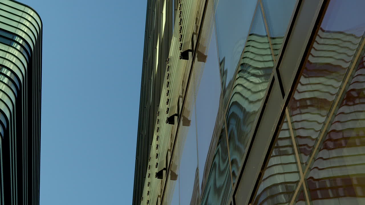 Close-up, angled view of a building's reflective glass facade, showing the distorted reflection of another building and the sky