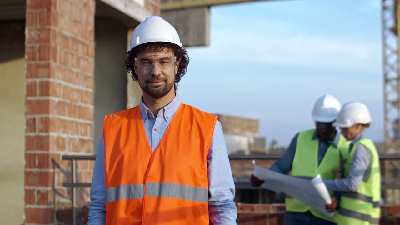retrato del apuesto hombre caucásico con casco y gafas sonriendo y posando para la cámara en el sitio de construcción. dos colegas multiétnicos, hombre y mujer hablando sobre el trabajo en el fondo.