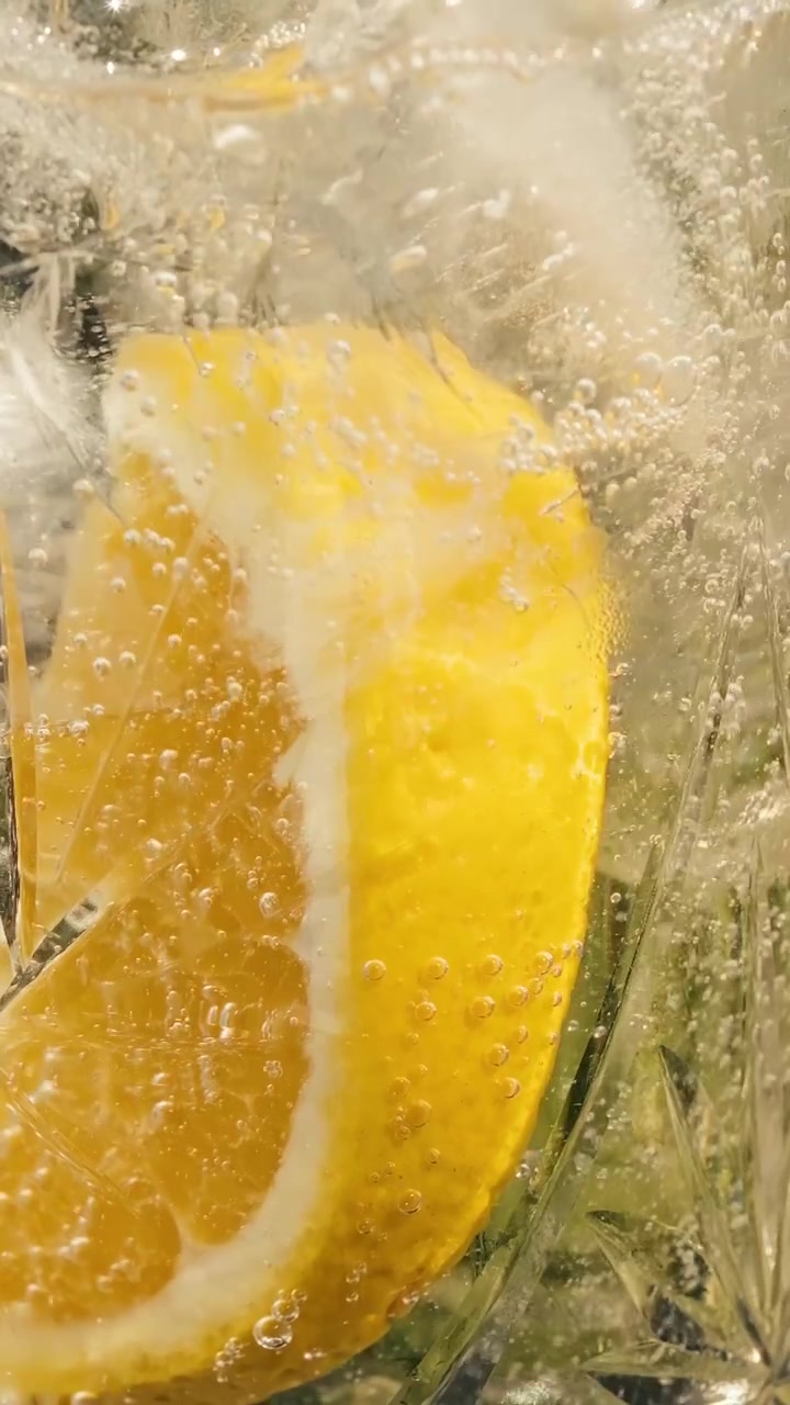 Cinematic vertical macro close-up of a refreshing cocktail with an orange slice and ice. A carbonated drink is poured, causing sparkling bubbles to swirl and dance dynamically around the ingredients