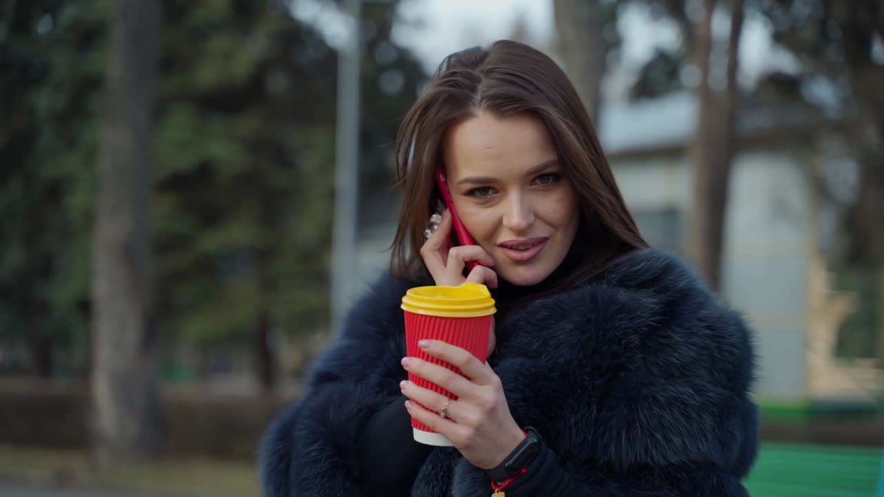 Attractive girl outdoors. Young woman in fur coat talking the phone while drinking coffee in the park. Portrait of a female with plastic cup. Slow motion.