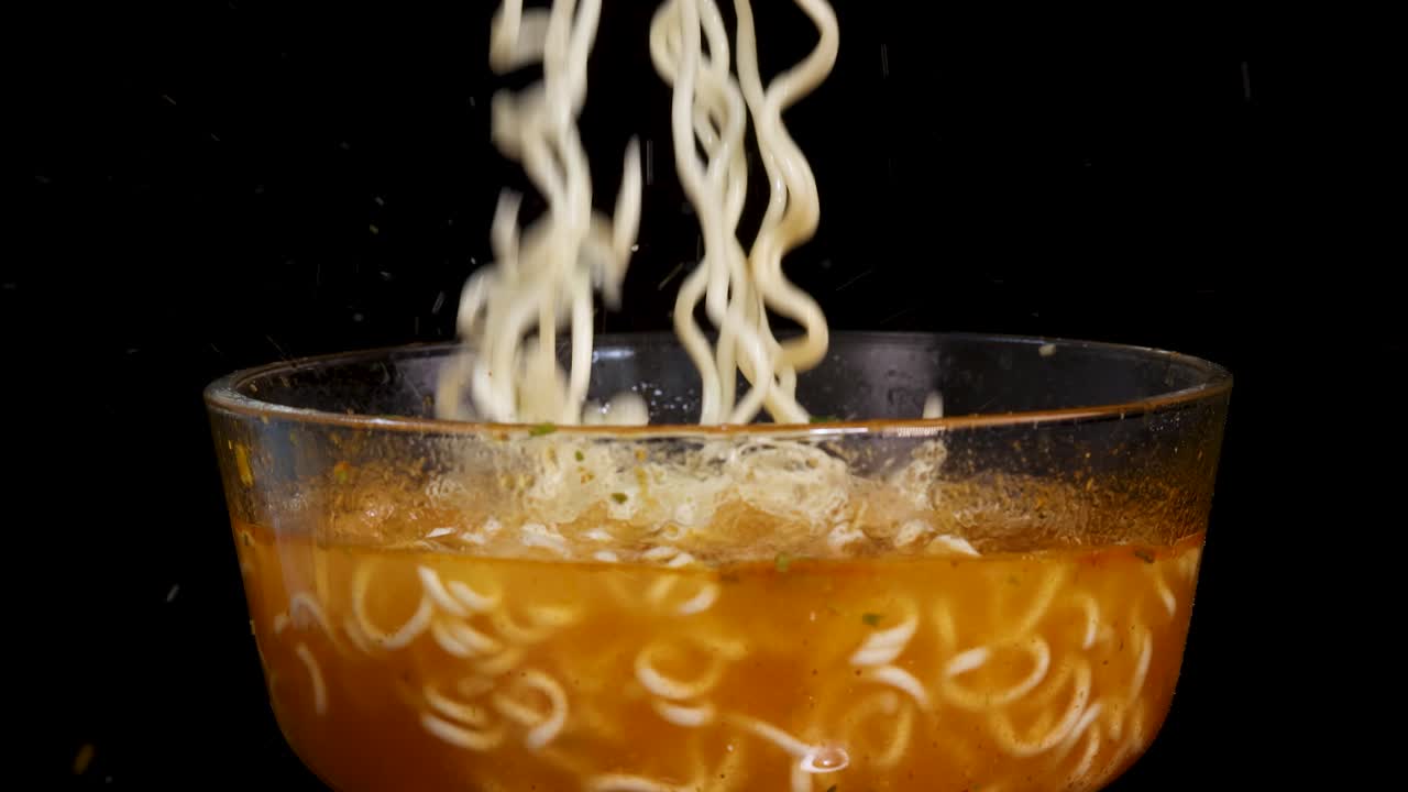 Cooked instant noodles fall into a steaming glass bowl of broth against a black background, captured in slow motion with dramatic studio lighting and a static camera