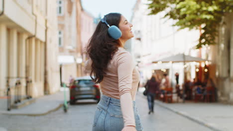 Happy-Indian-woman-in-wireless-headphones-listening-music-in-smartphone-app-dancing-on-city-street