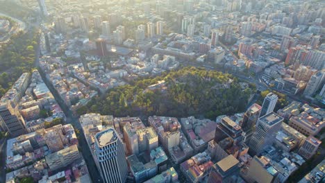 Drone-Aéreo-De-Arriba-Hacia-Abajo-Colina-Santa-Lucía-Santiago-De-Chile-Ciudad-Sol-Panorámico-Brillando-Sobre-La-Capital-Sudamericana,-Edificios-Calles-Parque-Urbano-A-La-Luz-Del-Día