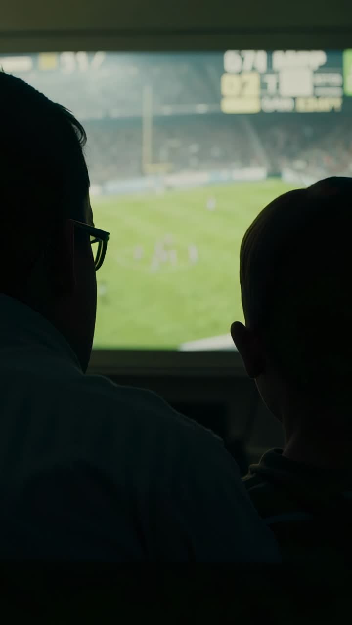Vertical video: Watching dad and child following score updates in suite on big screen, glasses seen