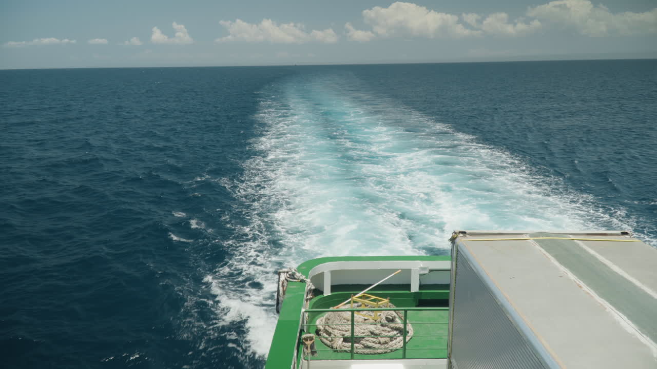 Fähre mit grünem Deck, die eine schaumige Kielwelle hinterlässt, Überfahrt auf offener See mit türkisfarbener Spur und Deckausrüstung, klarer Horizont und vereinzelte Wolken, rasche Fahrt und die Dynamik einer Küstenroute, kommerzielle Schifffahrtsatmosphäre.