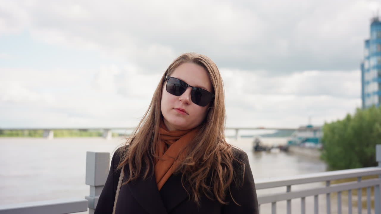 Young lady in dark coat and sunglasses standing near river railing under cloudy sky, modern buildings and bridge in background, calm wind and soft daylight create confident and stylish urban mood