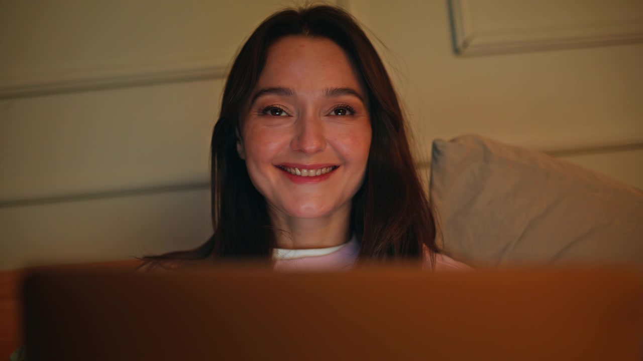 Brunetta che naviga contenuti online con il laptop seduta sul letto di notte, primo piano. Donna sorridente.