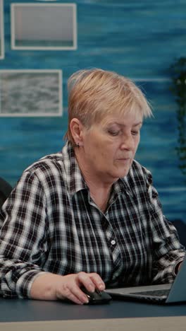Vertical video: Mature lady working on laptop at home workplace drinking coffee
