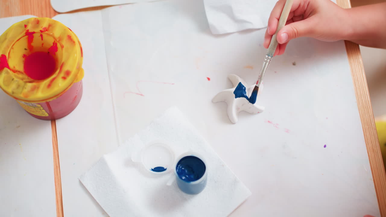 Close up of toddler hand holding paintbrush carefully applying blue paint on white star figurine while another hand reaches for brush nearby, art supplies scattered across table