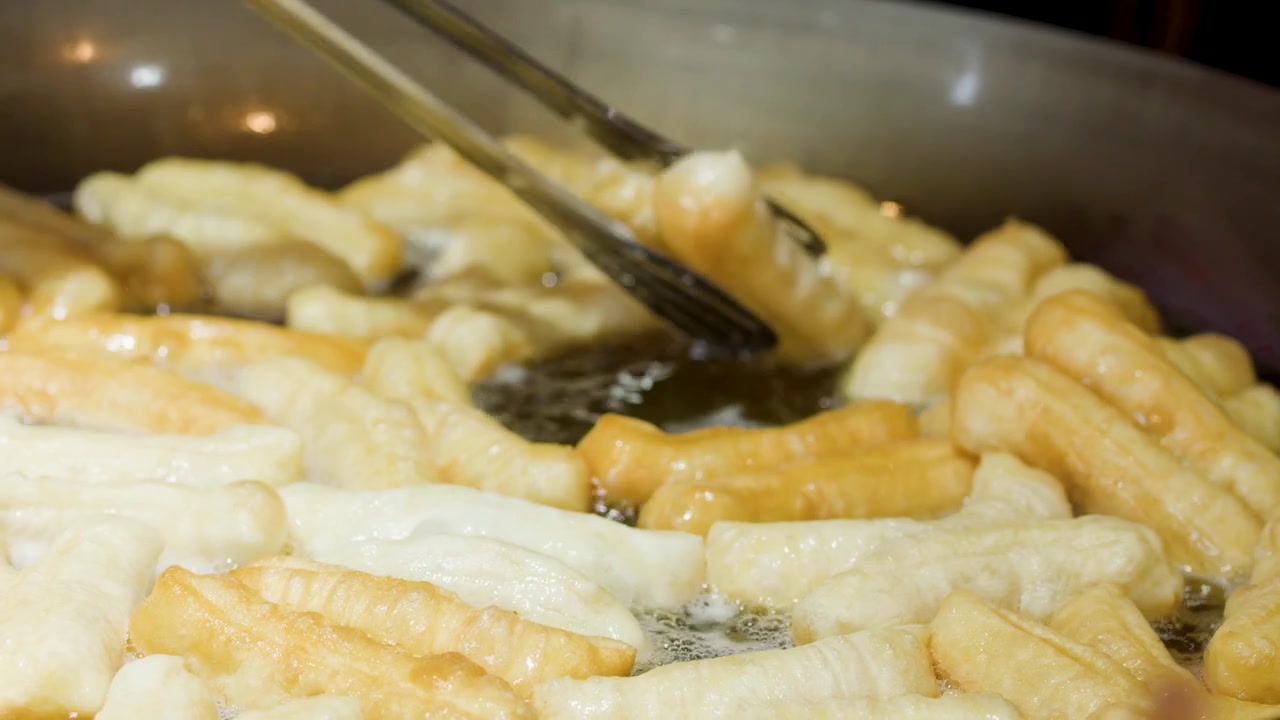 Primo piano di pinze di metallo che rigirano bastoncini di pasta fritta dorati in un wok gorgogliante.