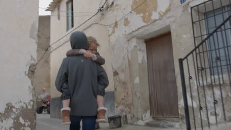 a woman carrying a child on her back walks down a narrow street in a small town.