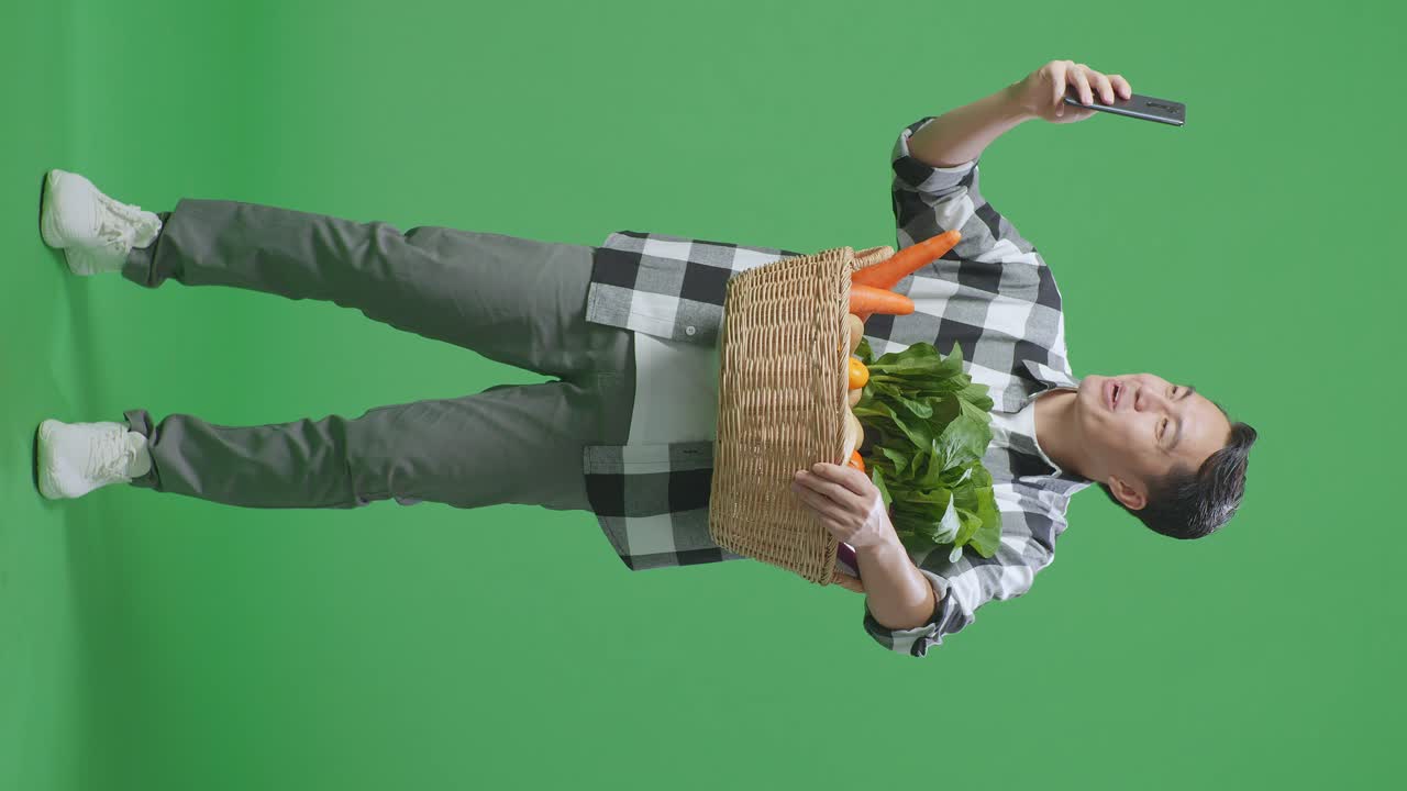 hombre sosteniendo una canasta de verduras, tomando una foto con un teléfono inteligente