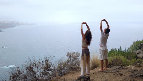 a man and a woman standing on the edge of a cliff overlooking the ocean raise their hands up and inhale the sea air during yoga