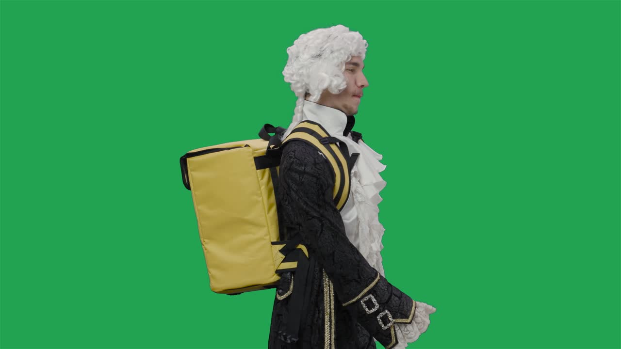 Portrait of courtier gentleman in black vintage suit and wig, walking with large yellow backpack fridge. Young man posing in studio with green screen background. Close up. Slow motion ready 59.94fps