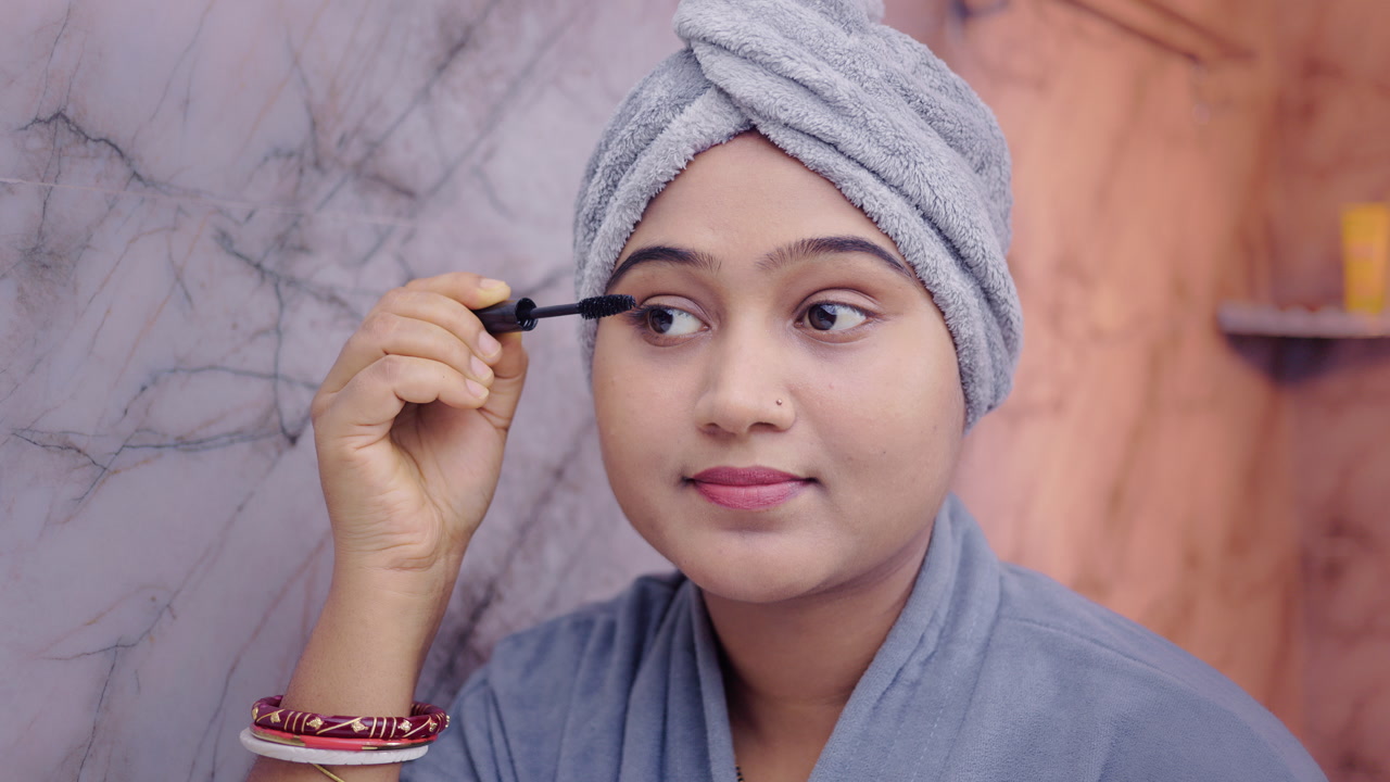 Indian woman applying mascara on eyelashes during her beauty routine, wearing bathrobe in the bathroom.