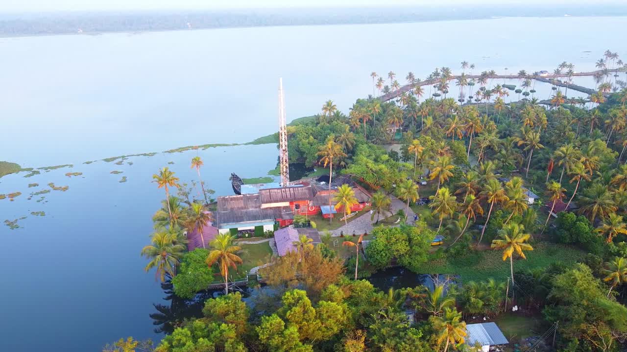 Gambar drone udara rumah tepi sungai yang menawan, tersembunyi di antara kebun kelapa yang rimbun dan tanaman hijau tropis di perairan belakang Kerala, saat matahari terbit yang tenang dan keemasan.
