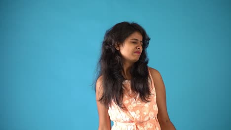 Ein-Junges-Indisches-Mädchen-In-Orangefarbenem-Kleid,-Das-Sich-Bemüht,-Hanteln-Zu-Heben,-Die-Auf-Einem-Isolierten-Blauen-Hintergrund-Stehen