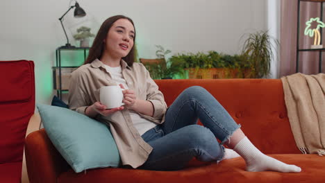 attractive smiling young woman drinking a cup of coffee or herbal tea relaxing resting at home couch