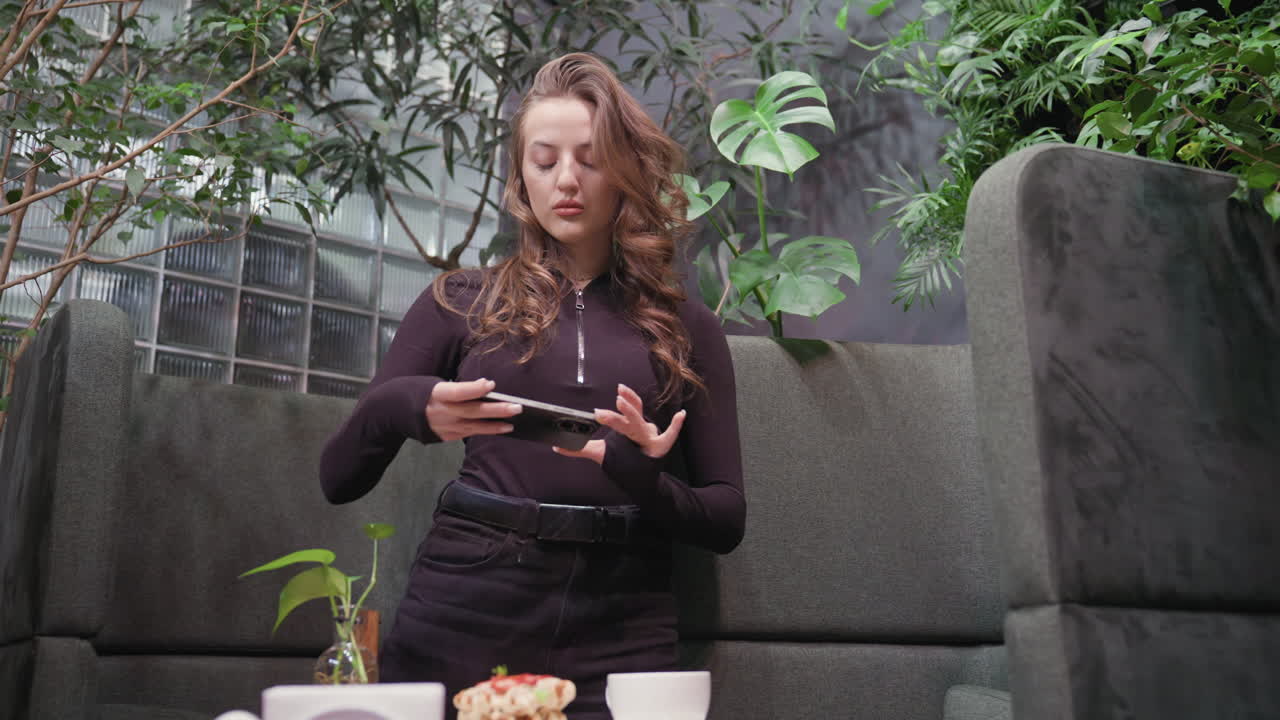 Lady in black top and trousers stands in leafy indoor environment holding phone in landscape mode while capturing overhead photo of waffles with toppings and latte on wooden table with cozy ambiance